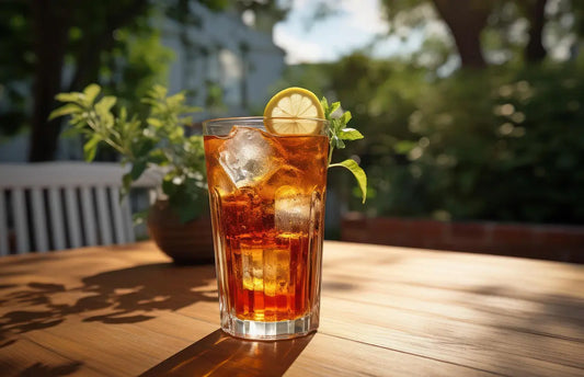 Verre de thĂ© glacĂ© dorĂ© posĂ© sur une table en bois baignĂ©e de soleil, garni de glaçons, dâagrumes et de feuilles de menthe, illustrant la fraĂźcheur estivale et la lĂ©gĂšretĂ© dâun moment dĂ©tente.